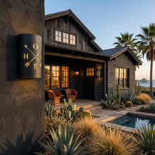 Load image into Gallery viewer, HOME and crossed-arrows wall sconce in black, mounted on a stucco column outside a timber and stone house