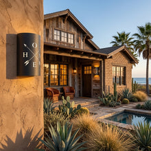Load image into Gallery viewer, HOME and crossed-arrows wall sconce in oil-rubbed bronze, mounted on a stucco column outside a timber and stone house