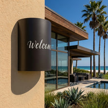 Load image into Gallery viewer, Welcome script wall sconce in oil-rubbed bronze, mounted on a stucco wall outside a modern beach house