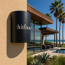 Load image into Gallery viewer, Welcome script wall sconce in black, mounted on a stucco wall outside a modern beach house